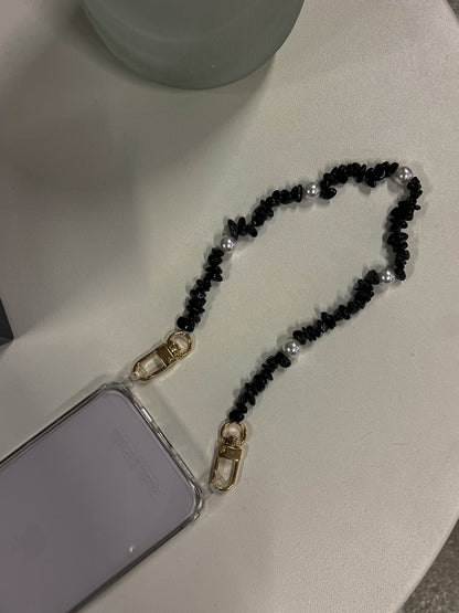Black beaded Shell Whisper SHORT PHONE STRAP attached to a clear phone case, styled on a white surface.