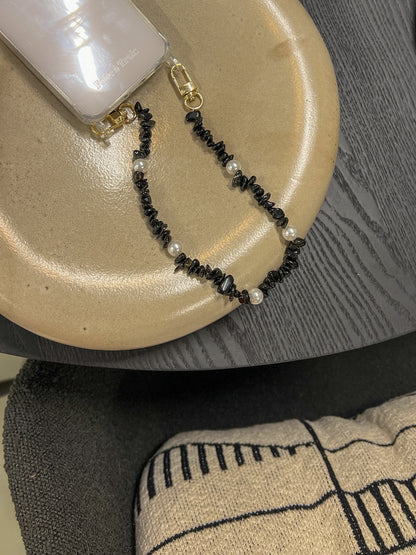 Black beaded Shell Whisper SHORT PHONE STRAP attached to a clear phone case, styled on a tan plate with dark textile background.