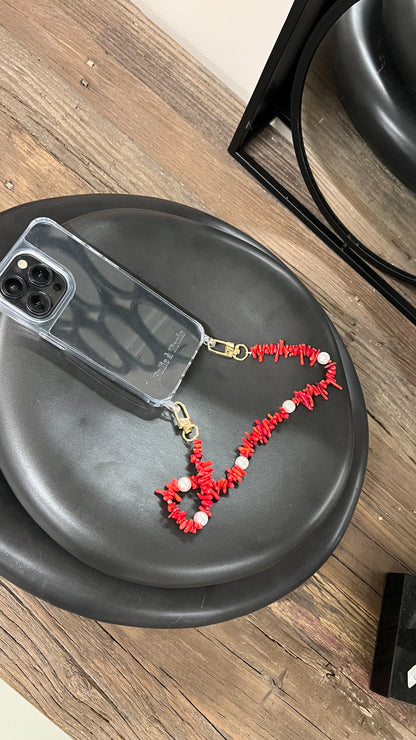 Red beaded Shell Whisper SHORT PHONE STRAP attached to a clear phone case, styled on a black stool on a wood floor.