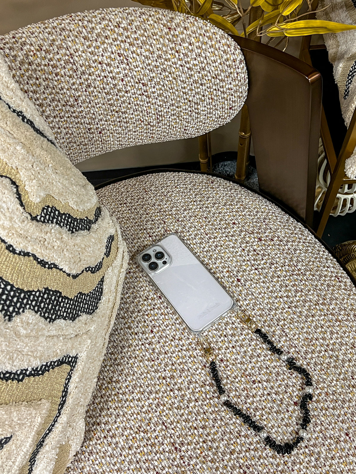 Black beaded Shell Whisper SHORT PHONE STRAP attached to a phone, resting on a tweed chair with gold decor in the background.