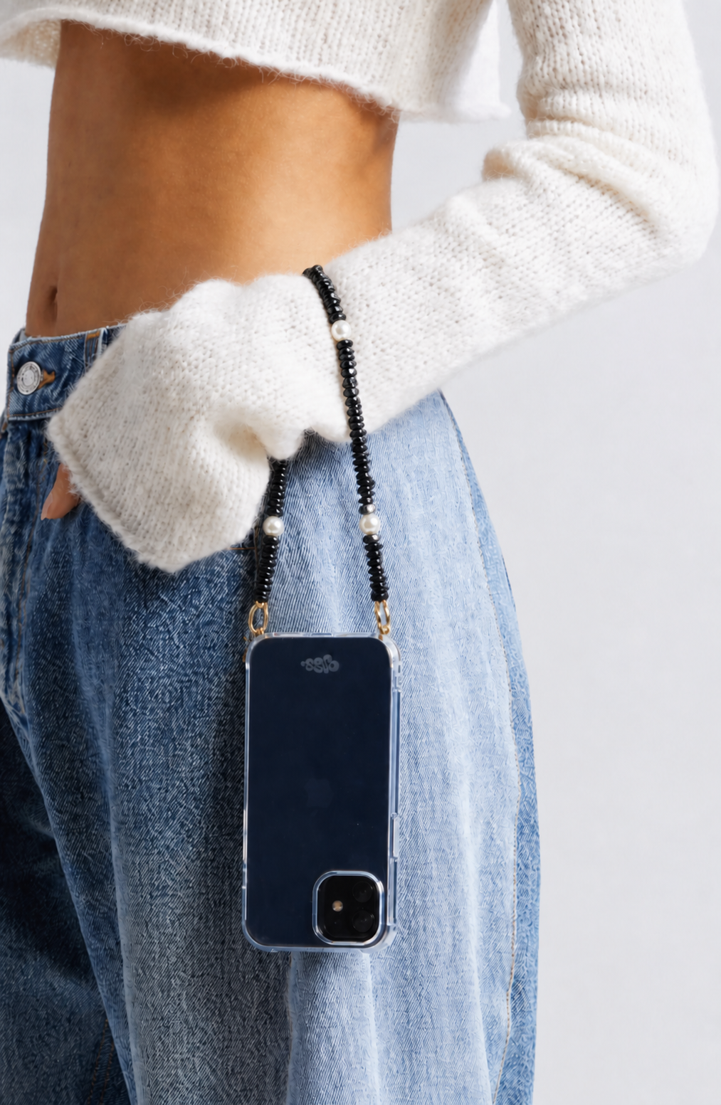 Black beaded Shell Whisper SHORT PHONE STRAP on a phone in a clear case, worn at the hip by a model in a cream sweater and jeans.