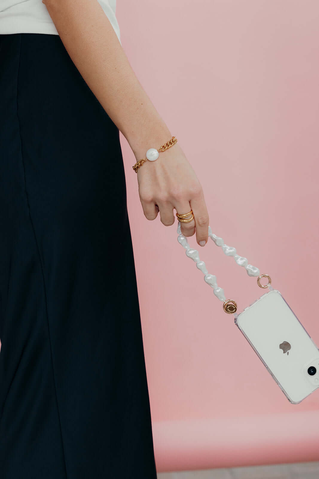 Woman holding her phone by a baroque pearl Short Pearl PHONE STRAP, wearing gold rings and a gold chain bracelet, pink background.