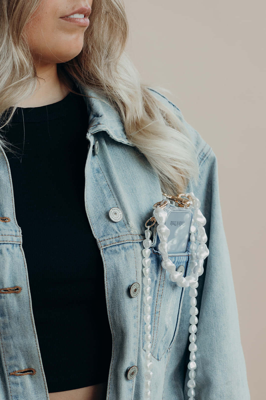 Phone with a baroque pearl Short Pearl PHONE STRAP tucked into a denim jacket pocket, close-up on the model's face and hair.