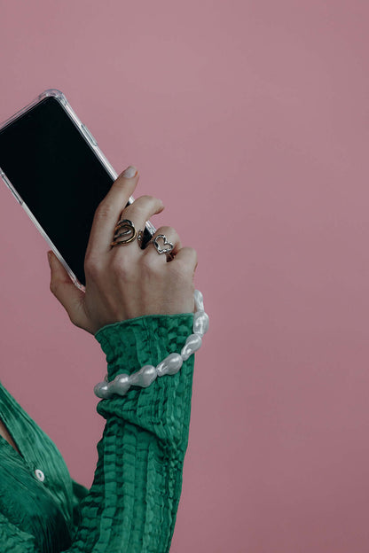 Hand wearing a baroque pearl Short Pearl PHONE STRAP around the wrist while holding a phone, green sleeve, pink background.
