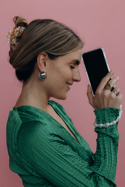 Woman in a green top talking on the phone with a baroque pearl Short Pearl PHONE STRAP visible on her wrist, pink background.