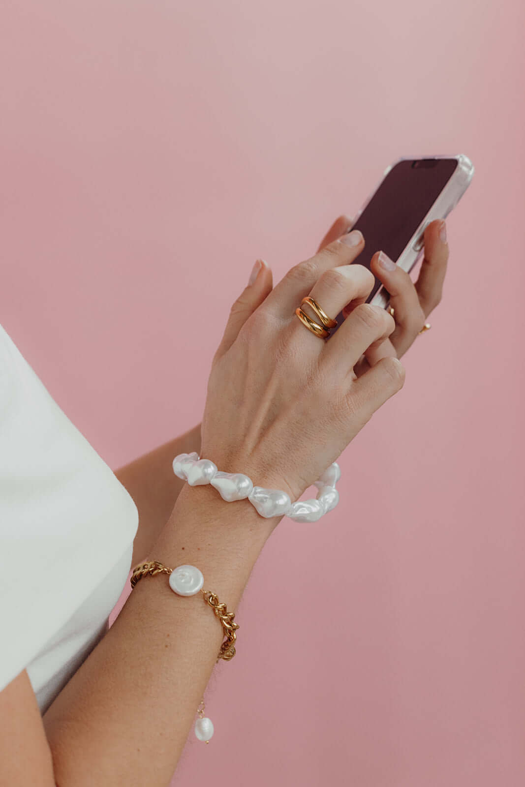 Hand with gold rings holding a phone, wearing a baroque pearl Short Pearl PHONE STRAP alongside a gold chain pearl-charm bracelet.