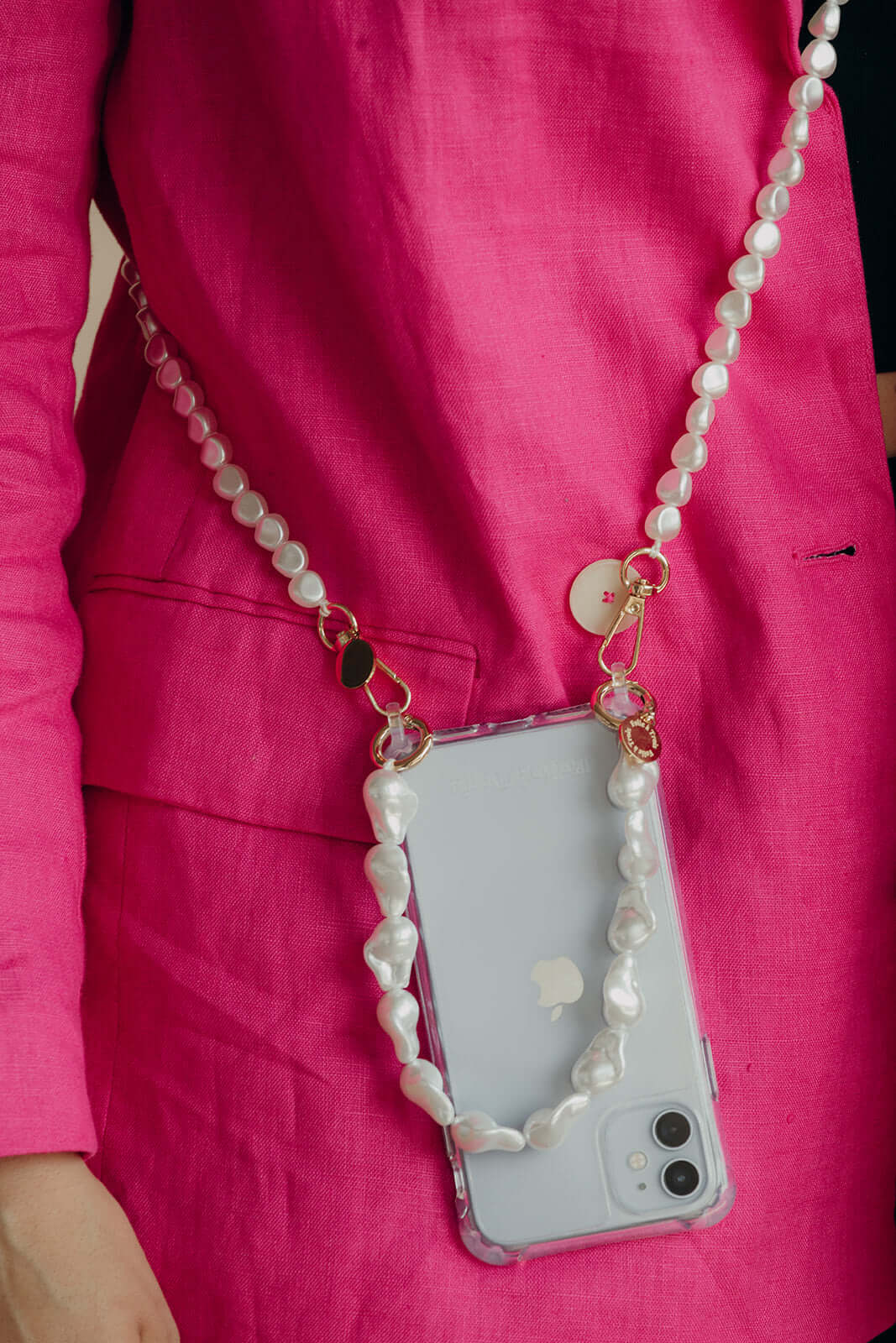 Long strand of baroque pearls worn draped around the neck and attached to a phone case, styled with a pink blazer outfit.