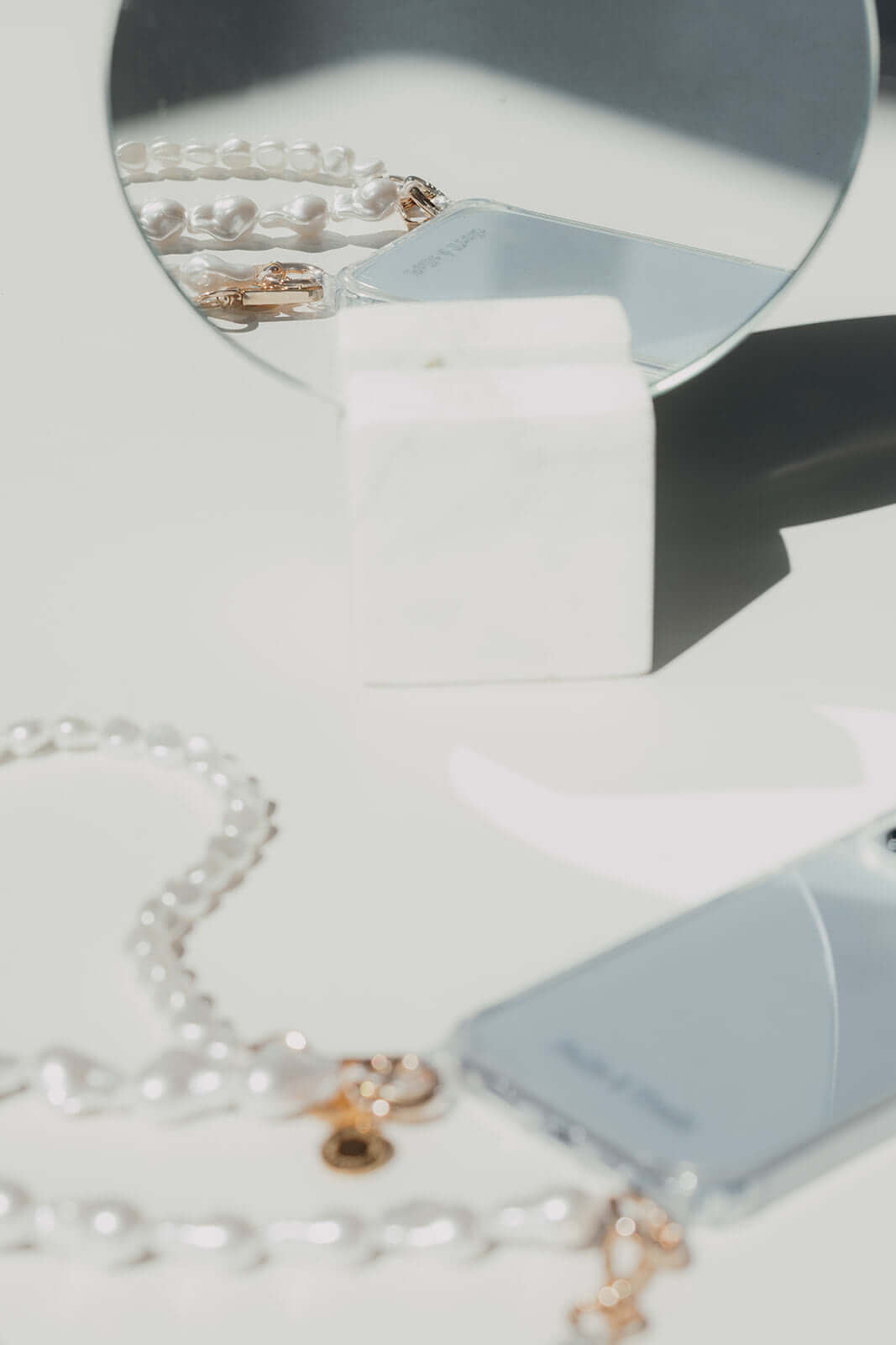 Blurred styled flat lay of a baroque pearl Short Pearl PHONE STRAP and phone beside a round mirror on a marble stand.