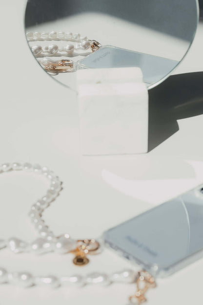 Blurred styled flat lay of a baroque pearl Short Pearl PHONE STRAP and phone beside a round mirror on a marble stand.