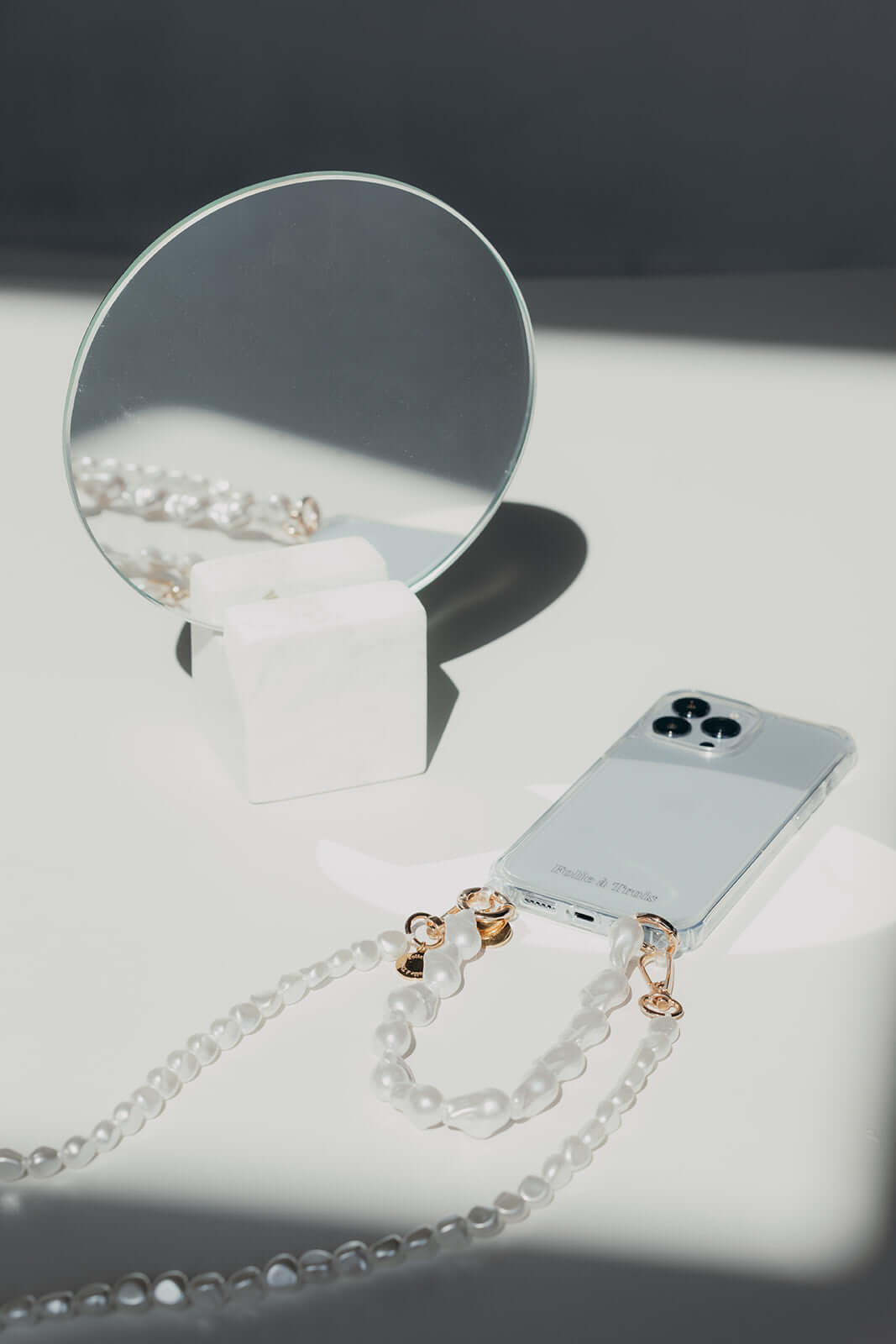 Baroque pearl Short Pearl PHONE STRAP and phone styled in sharp focus beside a round mirror on a marble stand.