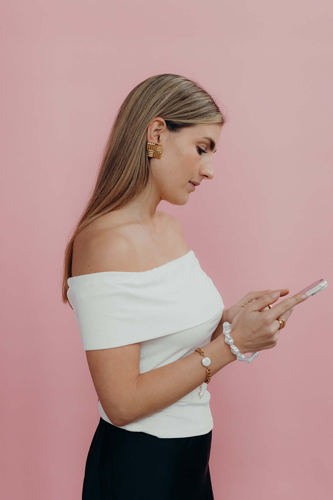 Woman in a white off-shoulder top texting, wearing a baroque pearl Short Pearl PHONE STRAP on her wrist, pink background.
