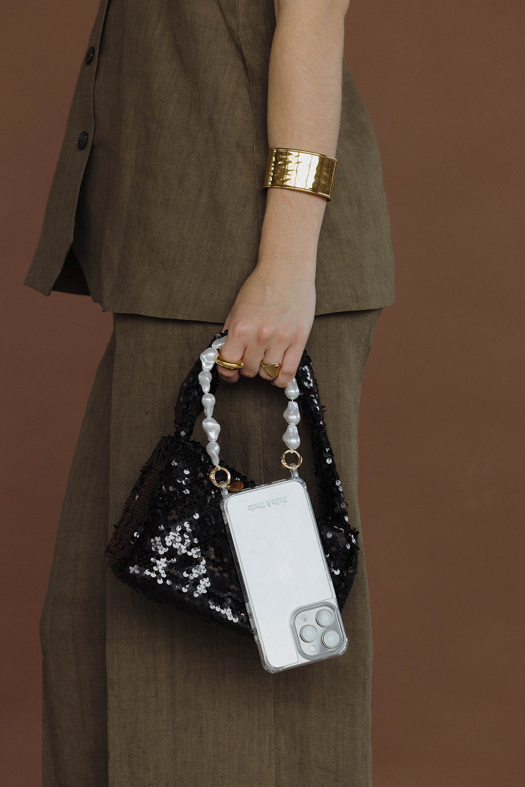 Woman holding a black sequin bag with a phone case attached by a baroque pearl Short Pearl PHONE STRAP, khaki suit.