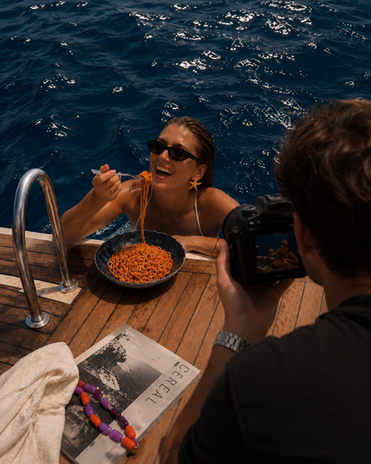 Wider shot of the Folie à Trois Siesta EARRINGS worn on a boat deck, with a photographer capturing the scene.