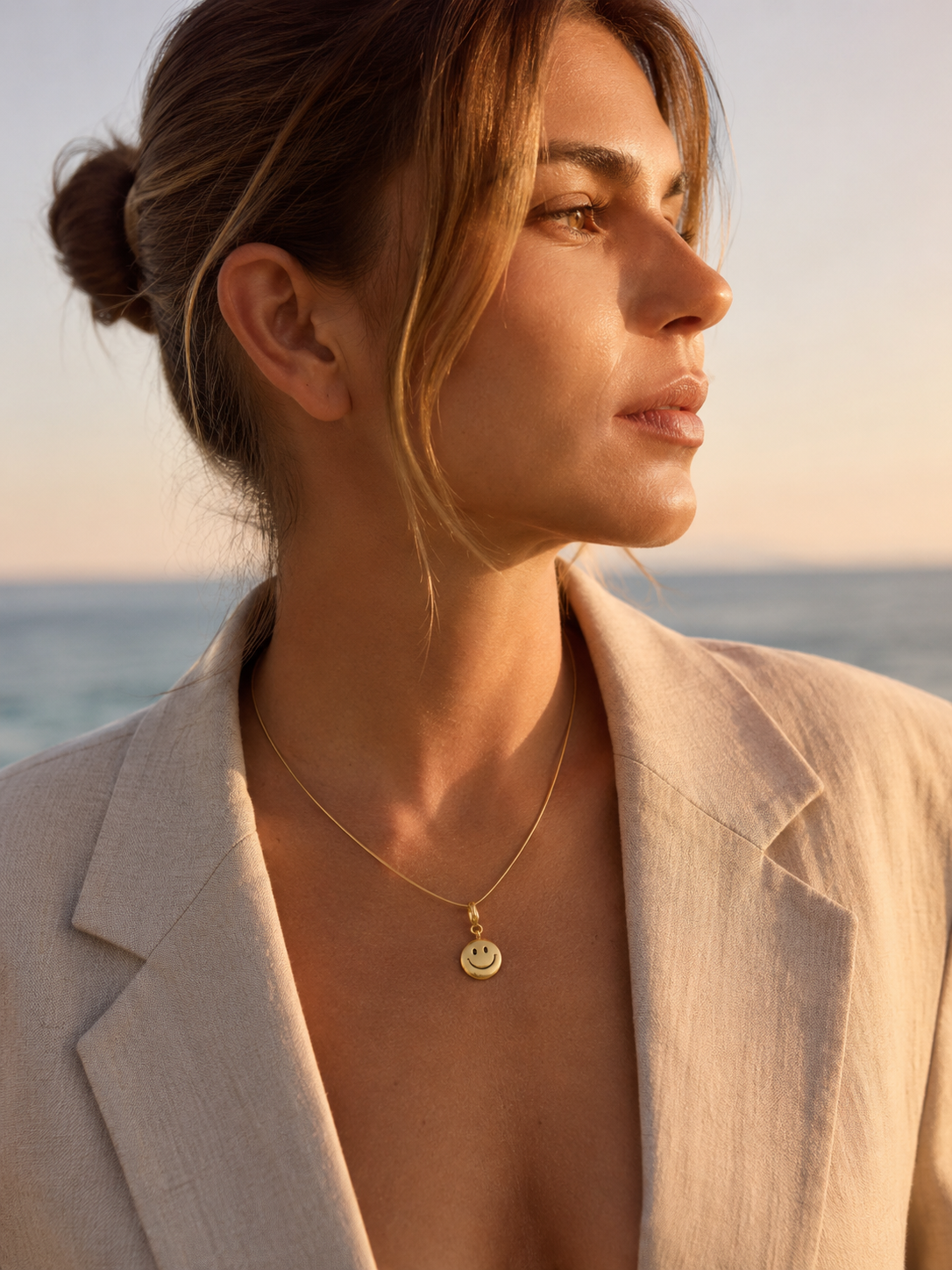 The Smiley Face CHARM worn as a pendant on a fine gold chain necklace, resting at the neckline at golden hour by the sea.