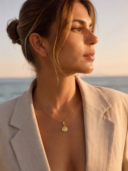 The Smiley Face CHARM worn as a pendant on a fine gold chain necklace, resting at the neckline at golden hour by the sea.