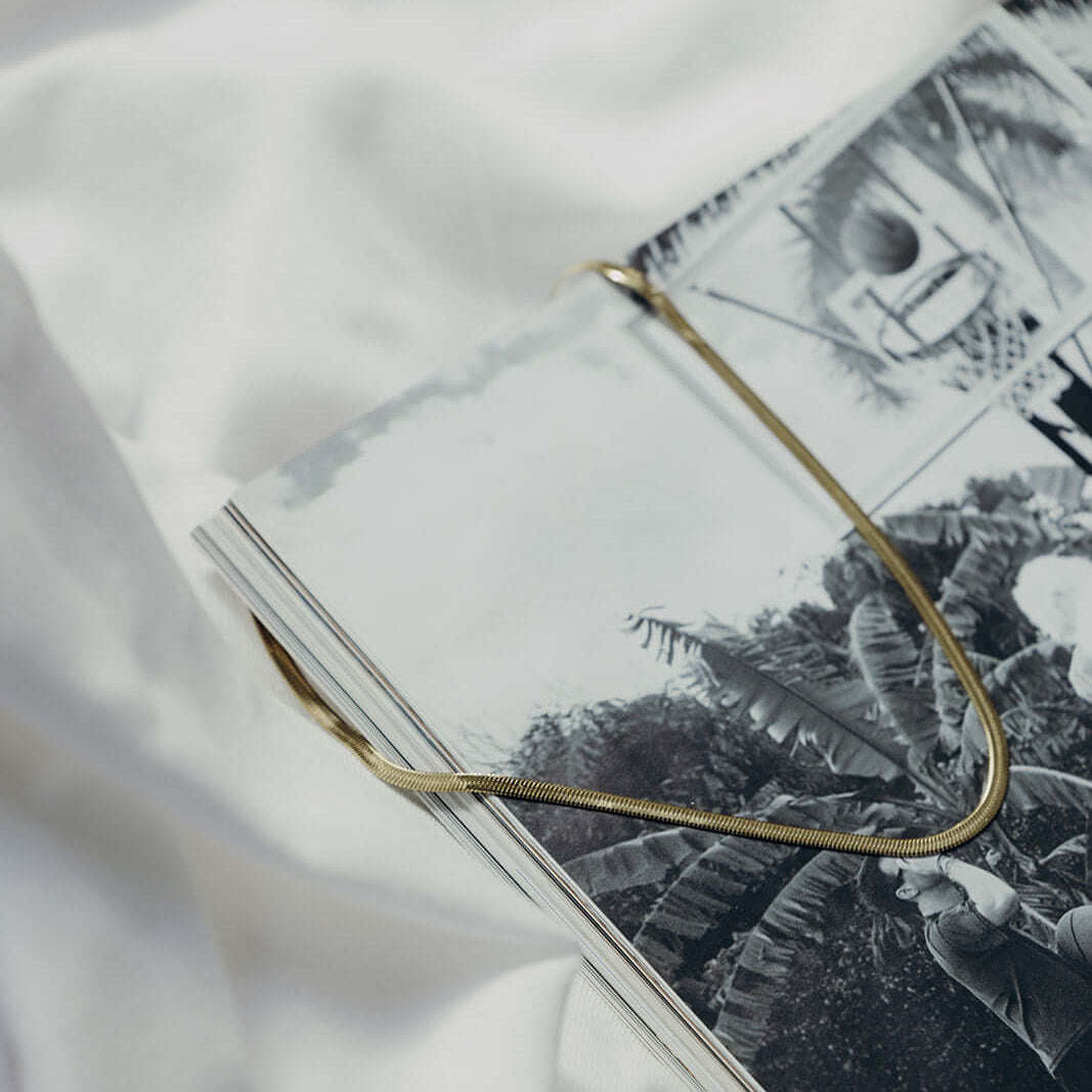 The Snake Skin NECKLACE in gold, the flat herringbone chain draped across an open magazine in an editorial still life.