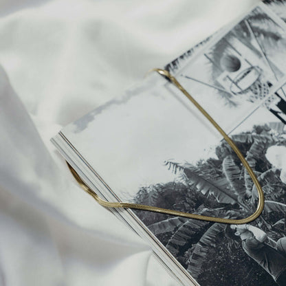 The Snake Skin NECKLACE in gold, the flat herringbone chain draped across an open magazine in an editorial still life.