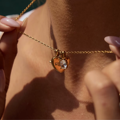 The Soft Power NECKLACE held between two fingers in extreme macro close-up, showing the engraved mom heart and crystal charm.