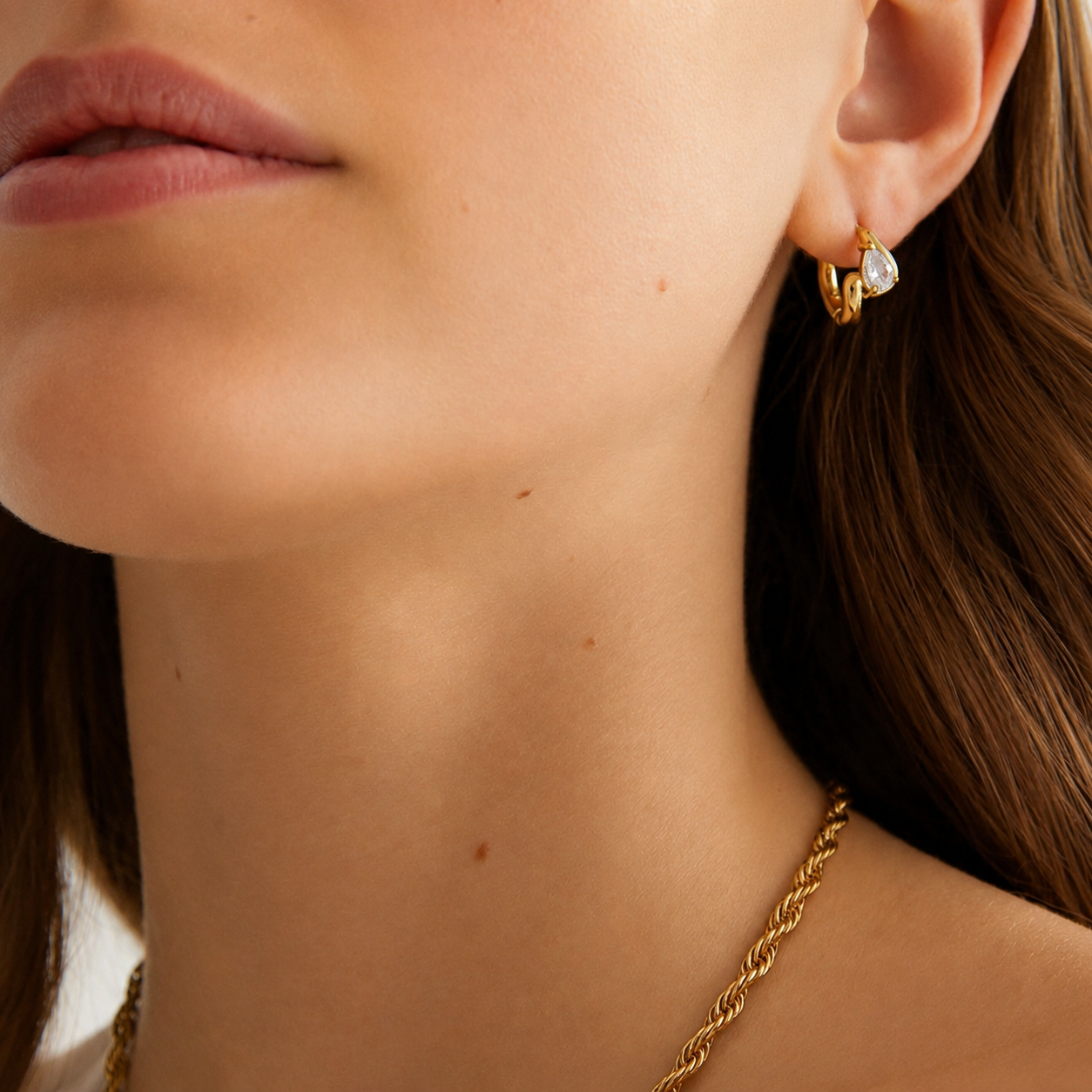 Model wears the Folie à Trois Sombra EARRINGS, a small gold huggie hoop with a pear-cut zirconia stone, layered with a rope chain necklace.