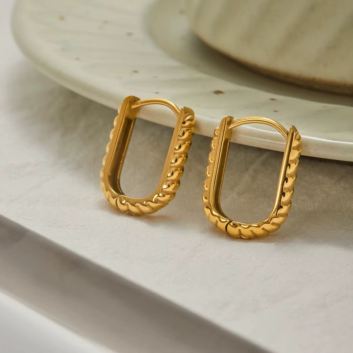 Close-up flat lay of the St. Moritz Earrings, a pair of ridged, textured gold oval hoops resting on a decorative plate.
