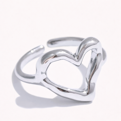 Open silver ring shaped like an outlined heart, shown at an angle on a cream backdrop — Folie à Trois Street Side RING.