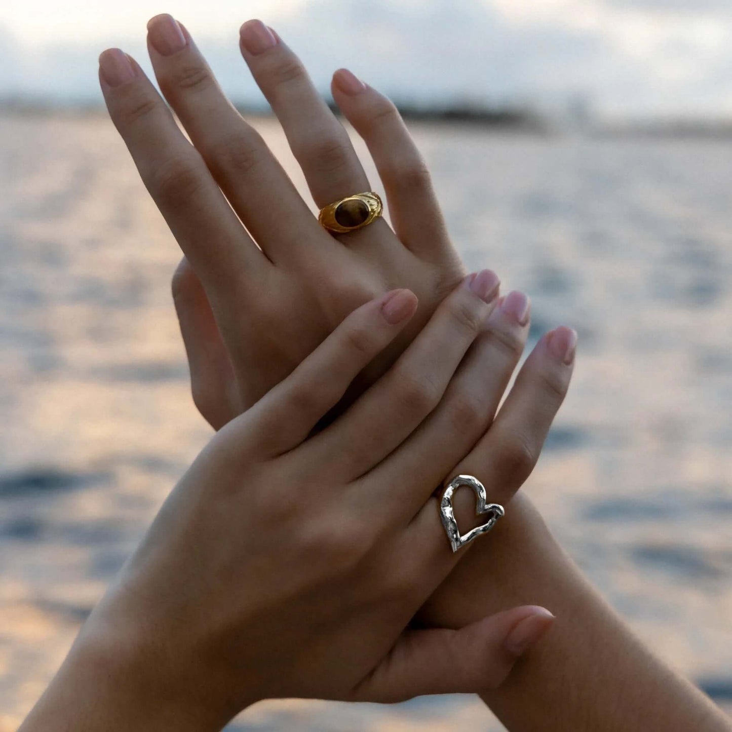Close-up of clasped hands near the ocean, one wearing the open silver heart-shaped Street Side RING, the other a gold dome ring.