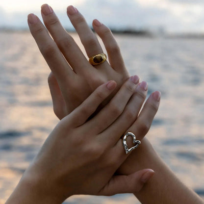 Close-up of clasped hands near the ocean, one wearing the open silver heart-shaped Street Side RING, the other a gold dome ring.