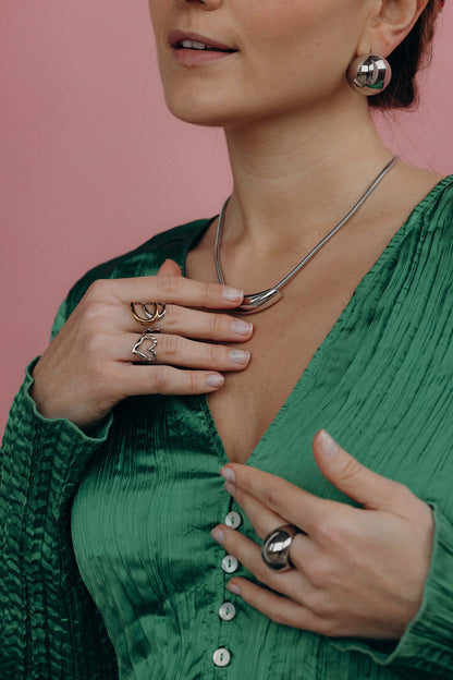 Close-up of a hand near the neck wearing the silver heart-shaped Street Side RING stacked with other rings, pink backdrop.
