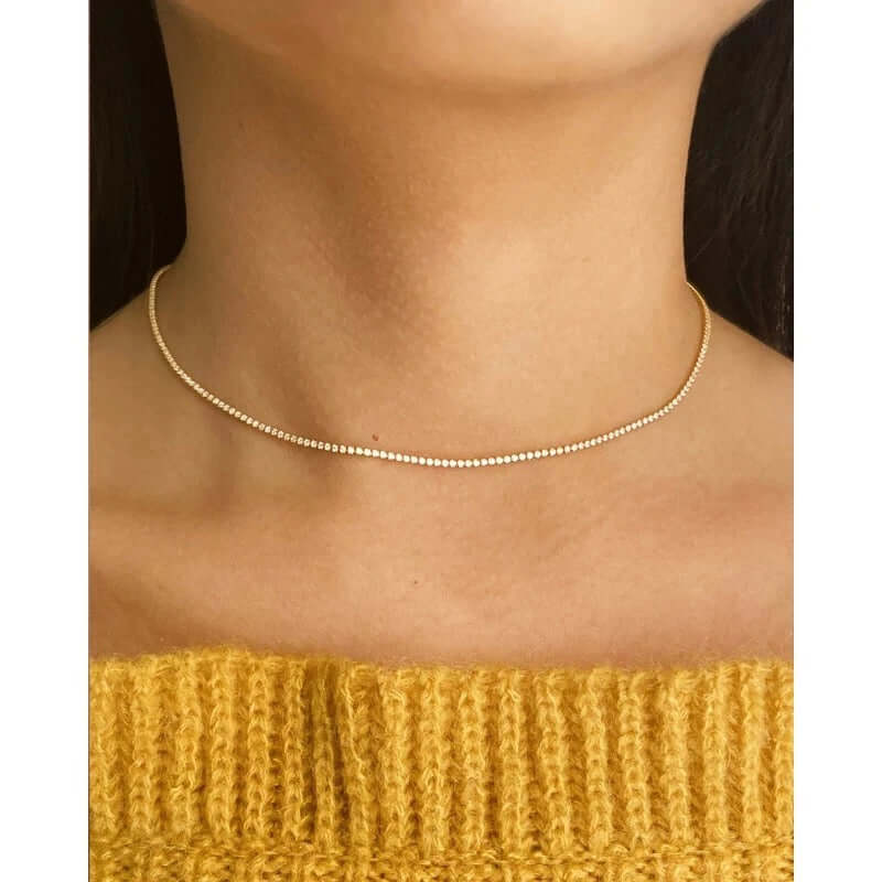 The Tennis NECKLACE in gold worn as a choker at the neckline, sparkling stones set close along the throat.