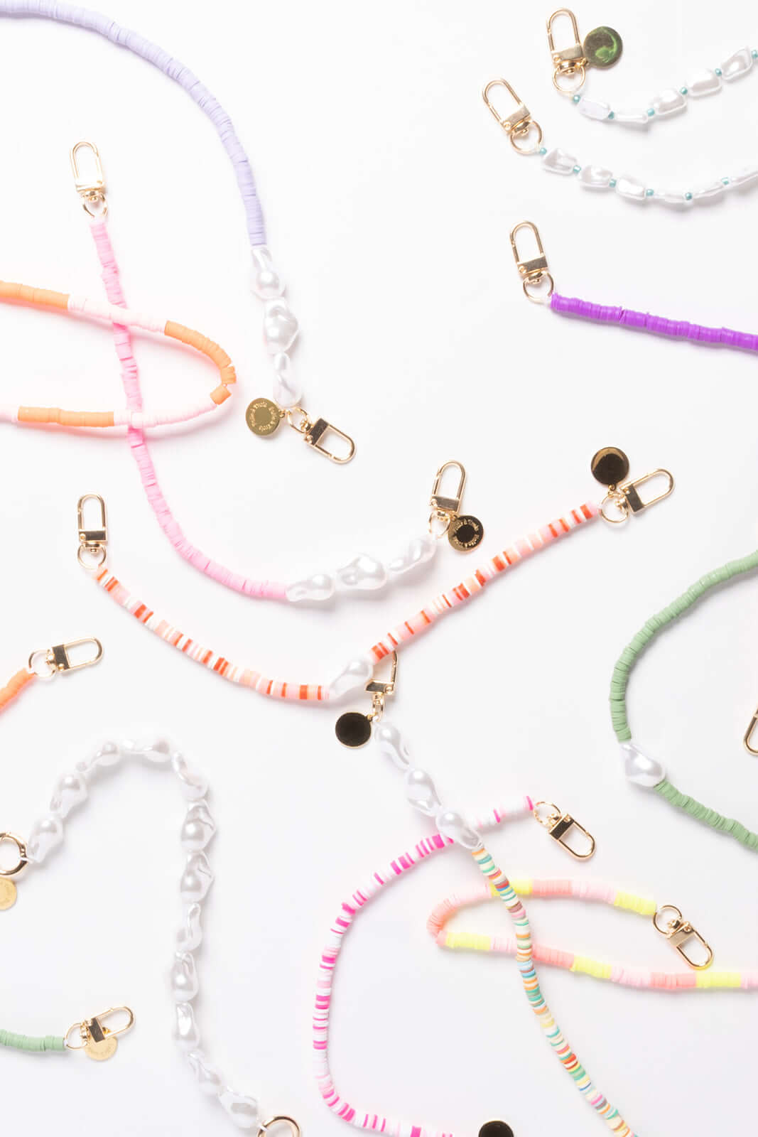 An assortment of heishi bead phone cords in lavender, pink, orange, purple, green, and pastel stripes, laid out with pearl beads and gold clasps.
