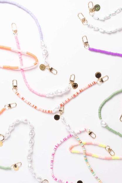 An assortment of heishi bead phone cords in lavender, pink, orange, purple, green, and pastel stripes, laid out with pearl beads and gold clasps.