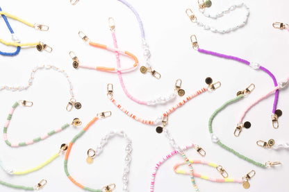 A close-up assortment of heishi bead phone cords in blue, yellow, lavender, pink, orange, purple, and green, laid out together with pearl beads.