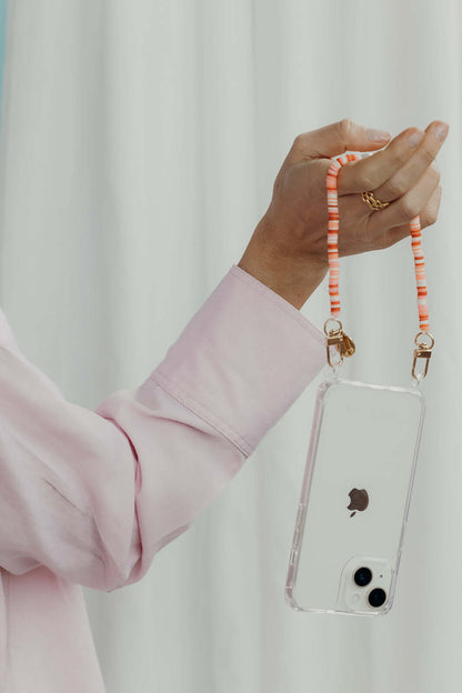 A hand holding a phone by its pink and orange striped heishi bead phone cord, against a white curtain backdrop.