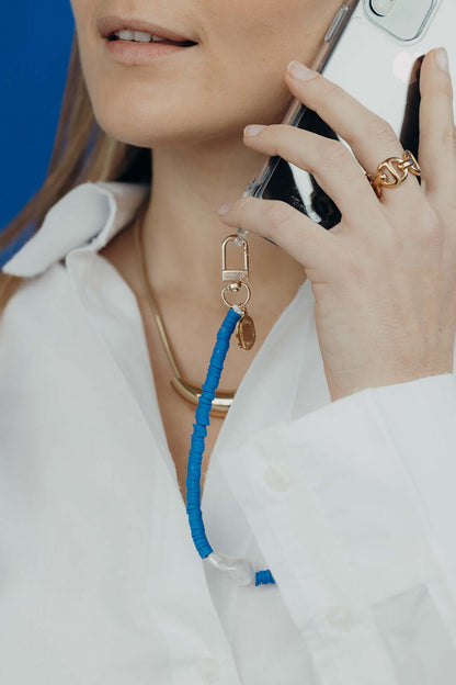 Close-up of a woman on the phone, wearing a blue heishi bead phone cord with a gold charm, over a white collared shirt.
