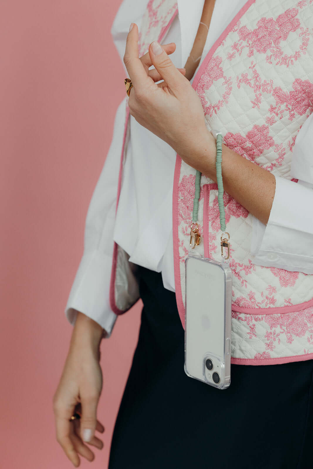 Close-up of a sage green heishi bead phone cord attached to a phone in a clear case, worn over a quilted pink and white vest.