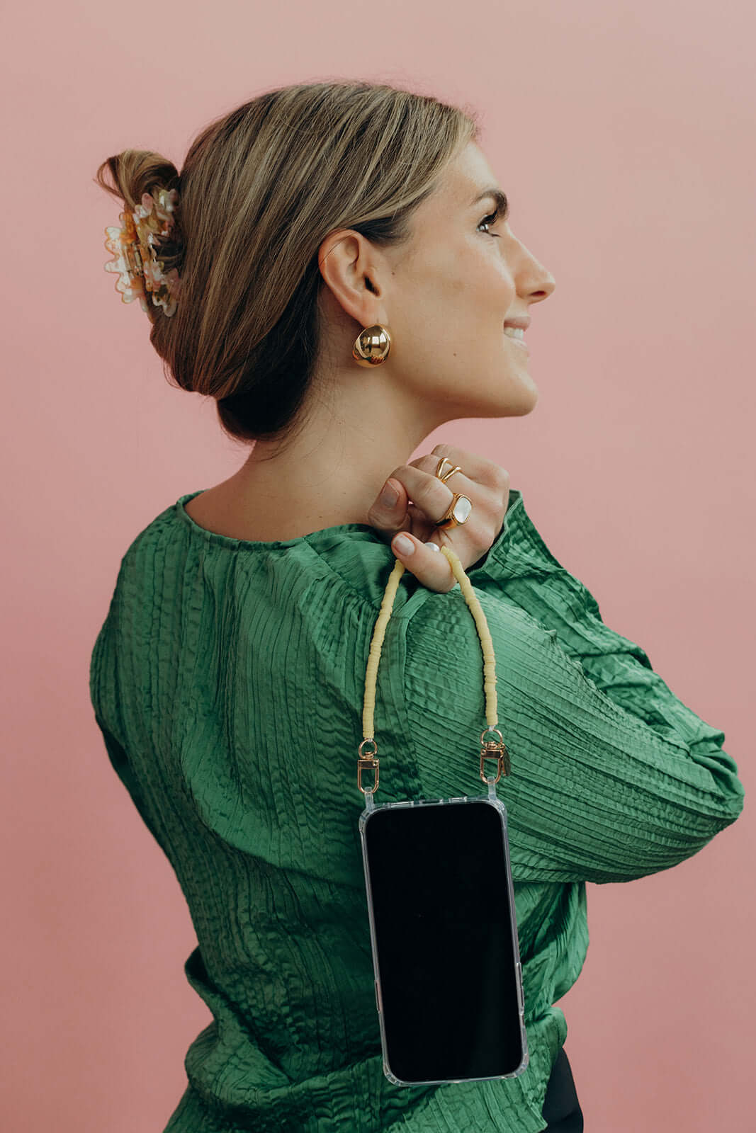 A woman looking over her shoulder, wearing a yellow and cream heishi bead phone cord with a phone, over a green blouse.