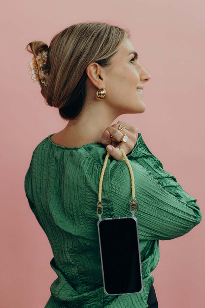 A woman looking over her shoulder, wearing a yellow and cream heishi bead phone cord with a phone, over a green blouse.