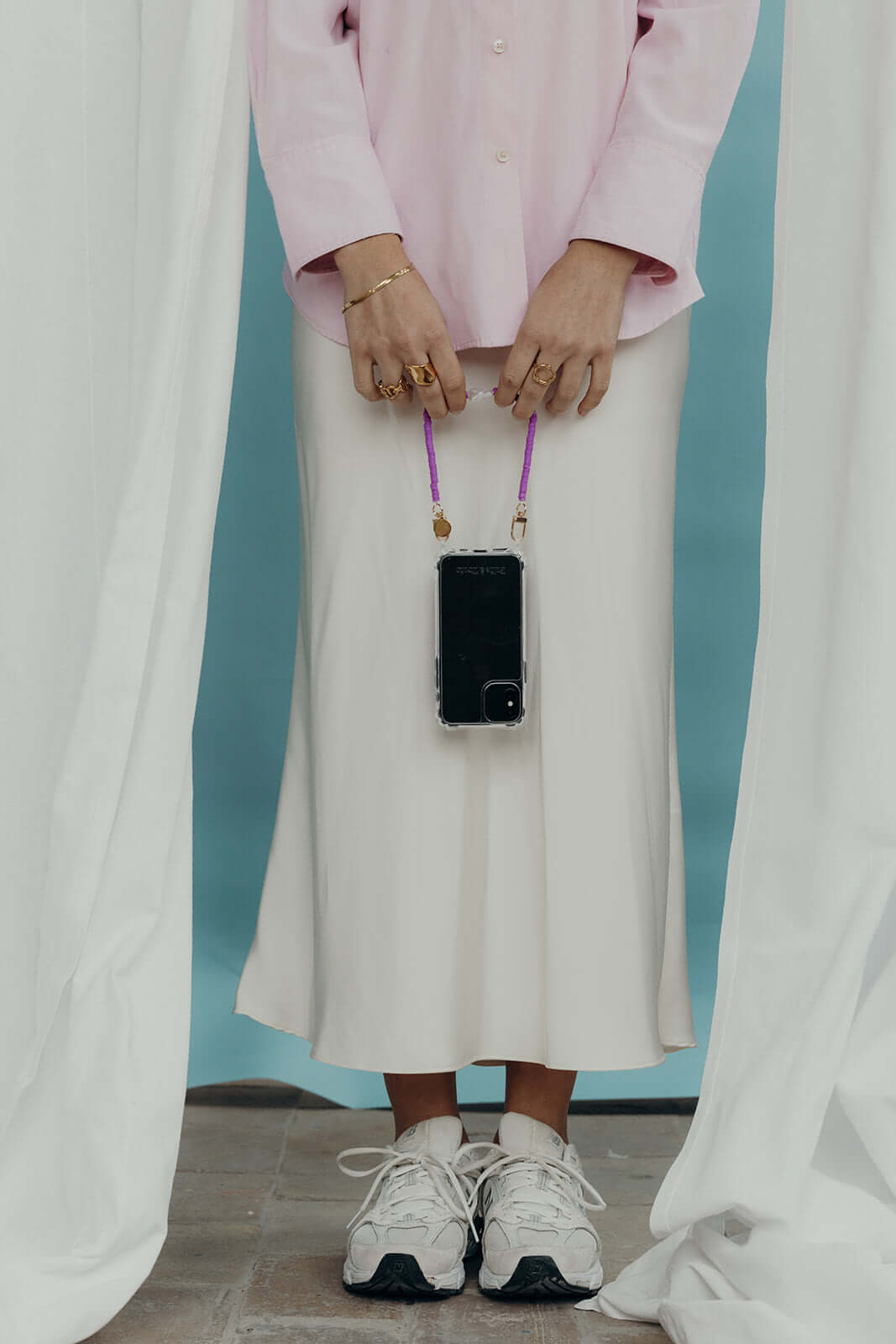 A woman holding a phone by its purple heishi bead cord, worn over a cream skirt and pink shirt, with white sneakers.