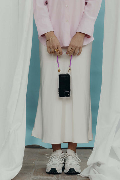 A woman holding a phone by its purple heishi bead cord, worn over a cream skirt and pink shirt, with white sneakers.