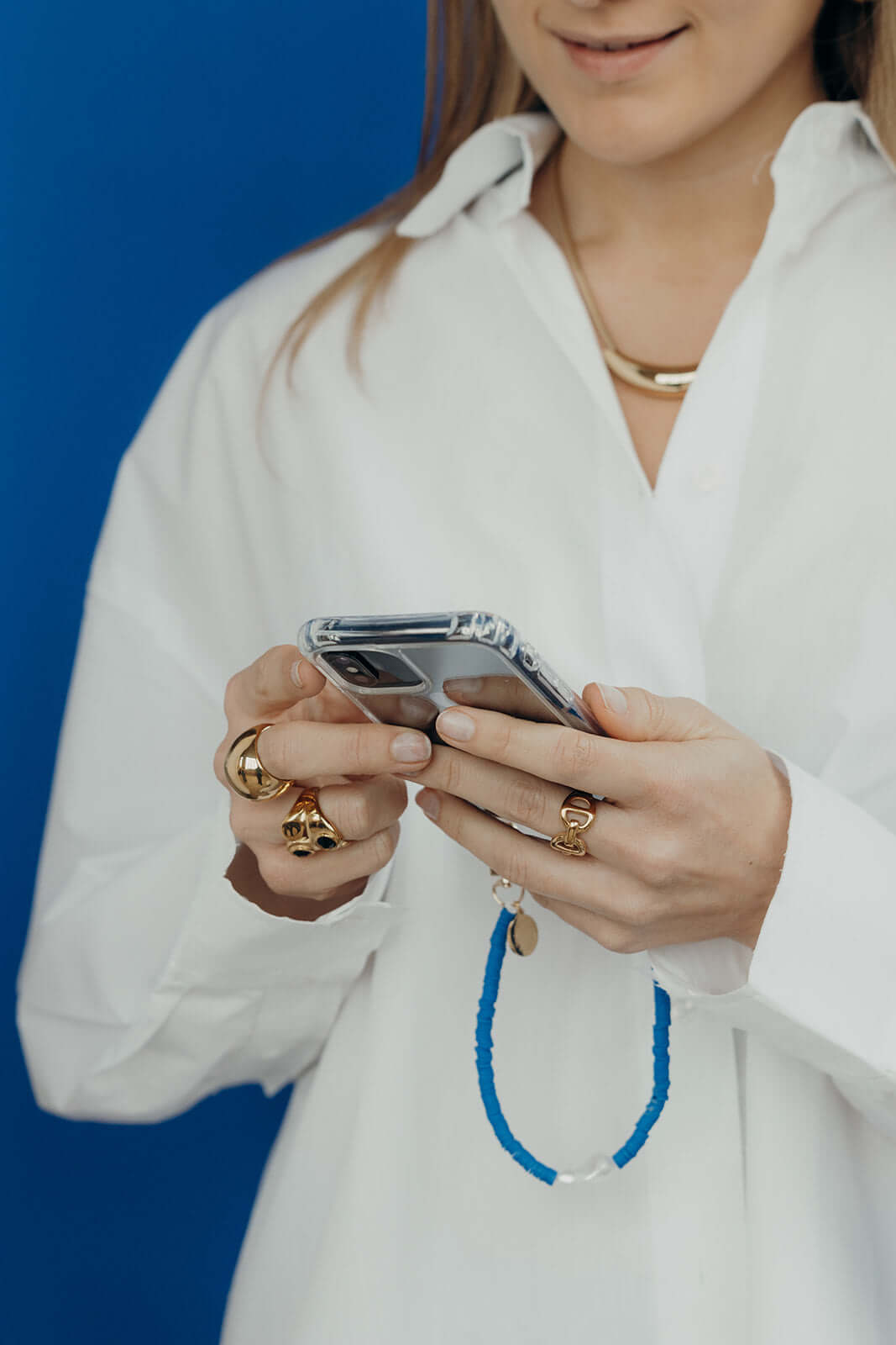 Close-up of a woman using her phone, wearing a blue and white heishi bead phone cord, over a white shirt against a blue background.