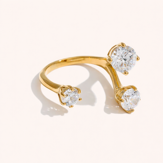 Open gold ring set with three round clear stones at different heights, shown at an angle on a cream backdrop — Trinity RING.