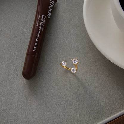 Open gold Trinity RING with three clear stones styled on a grey surface beside a lip tint tube and teacup.