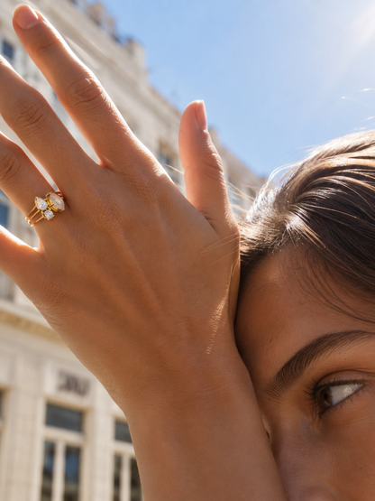 Model wears the gold Trio Halo RING stacked with other rings on her finger, hand raised near her face against a city street backdrop.