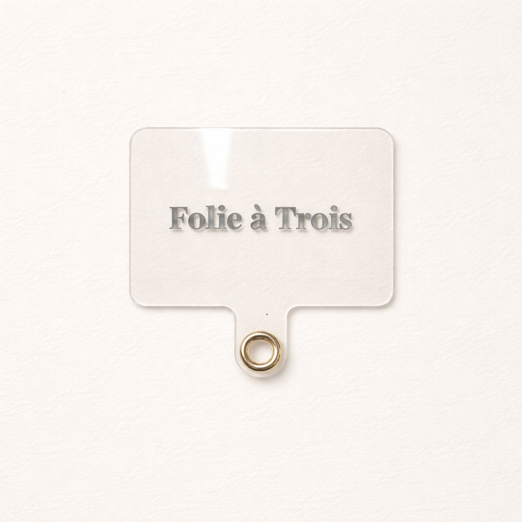 Close-up of a clear acrylic Folie à Trois tag with a gold grommet ring, the Universal Phone Adapter clip-on attachment.