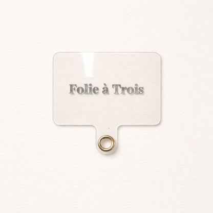 Close-up of a clear acrylic Folie à Trois tag with a gold grommet ring, the Universal Phone Adapter clip-on attachment.