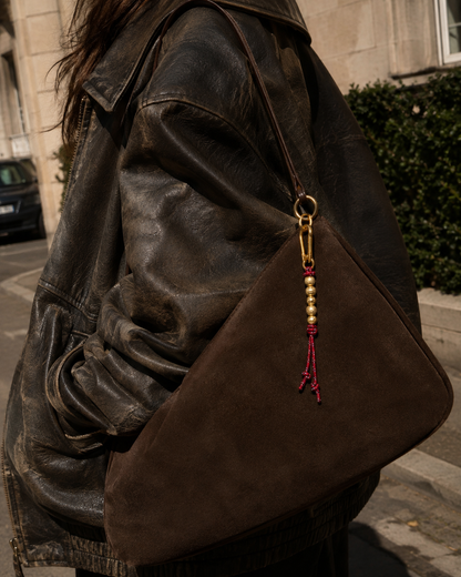 Folie à Trois Uva BAG CHARM, a red cord tassel with gold beads, clipped to a brown suede bag worn under a leather jacket.