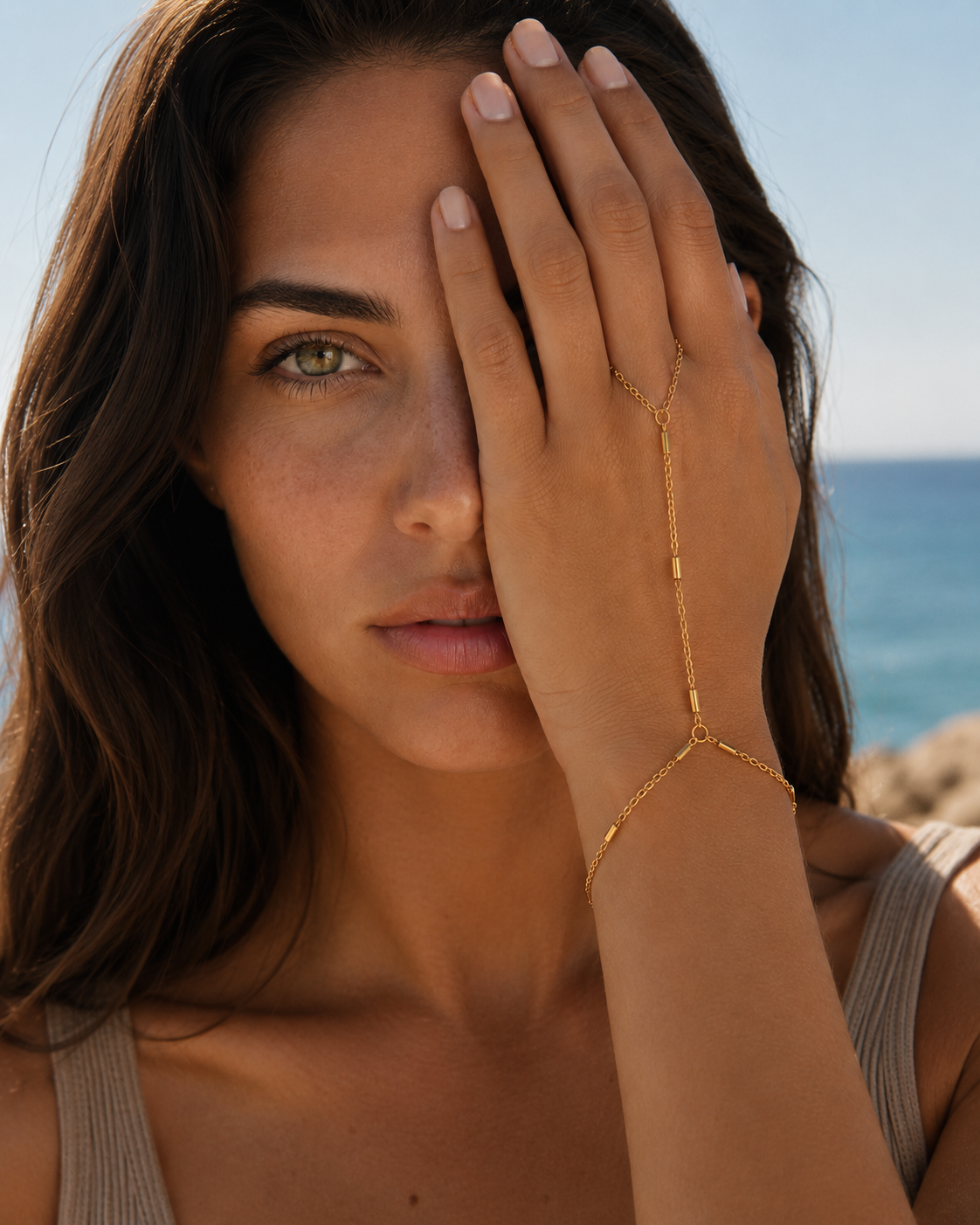 Venus Hand Chain BRACELET worn on the hand with the sea behind her, model covering one eye with her fingers.