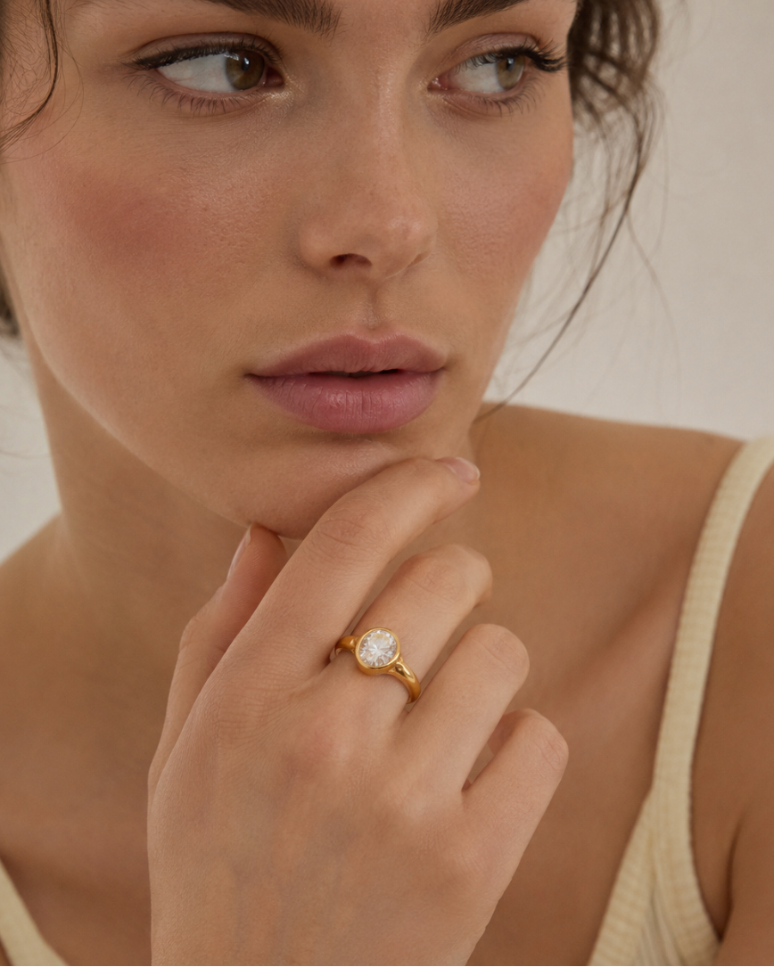 Model wears a gold Vertex RING bezel-set with an oval white zirconia stone, hand raised to the chin — Folie à Trois