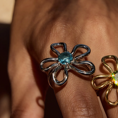 Extreme close-up of a hand wearing the silver wire-flower Vida RING with a blue stone center, gold version visible beside it.
