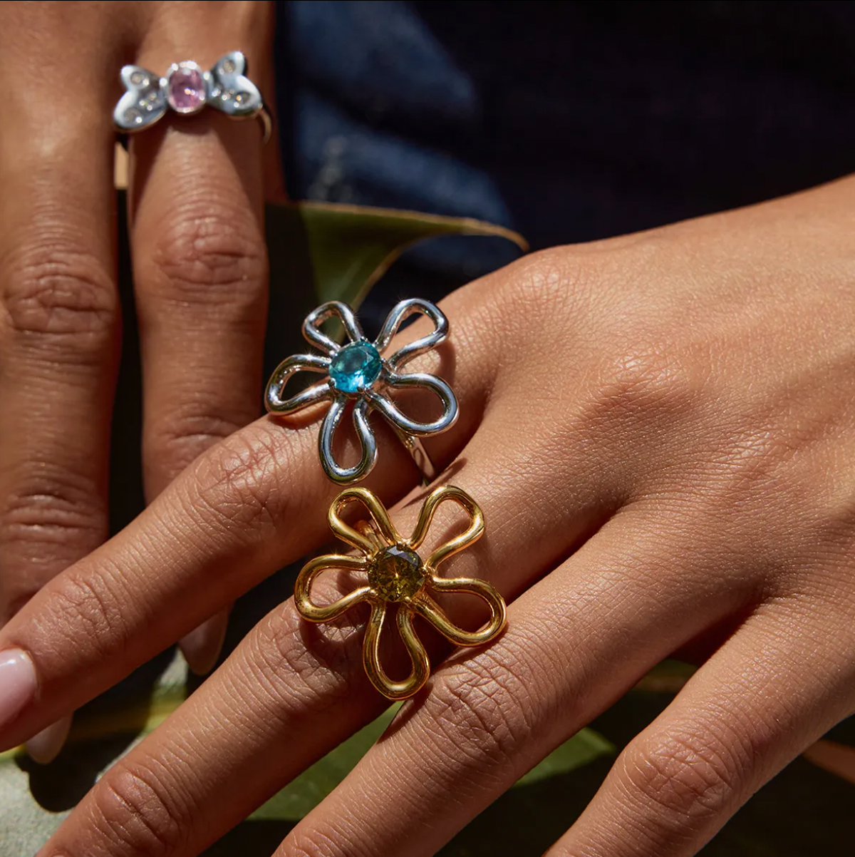 Close-up of a hand wearing the silver and gold wire-flower Vida RING styles stacked, each set with a different colored stone.